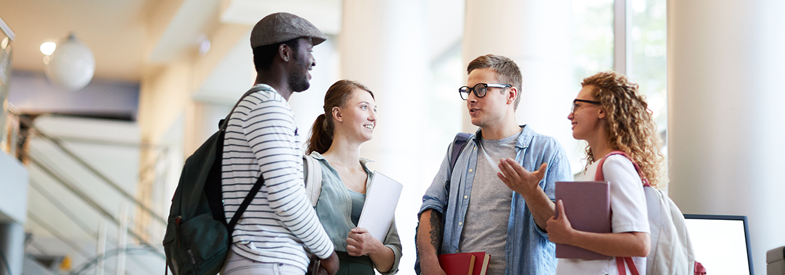 Image of four young adults in a lobby talking to each other and holding books and backpacks